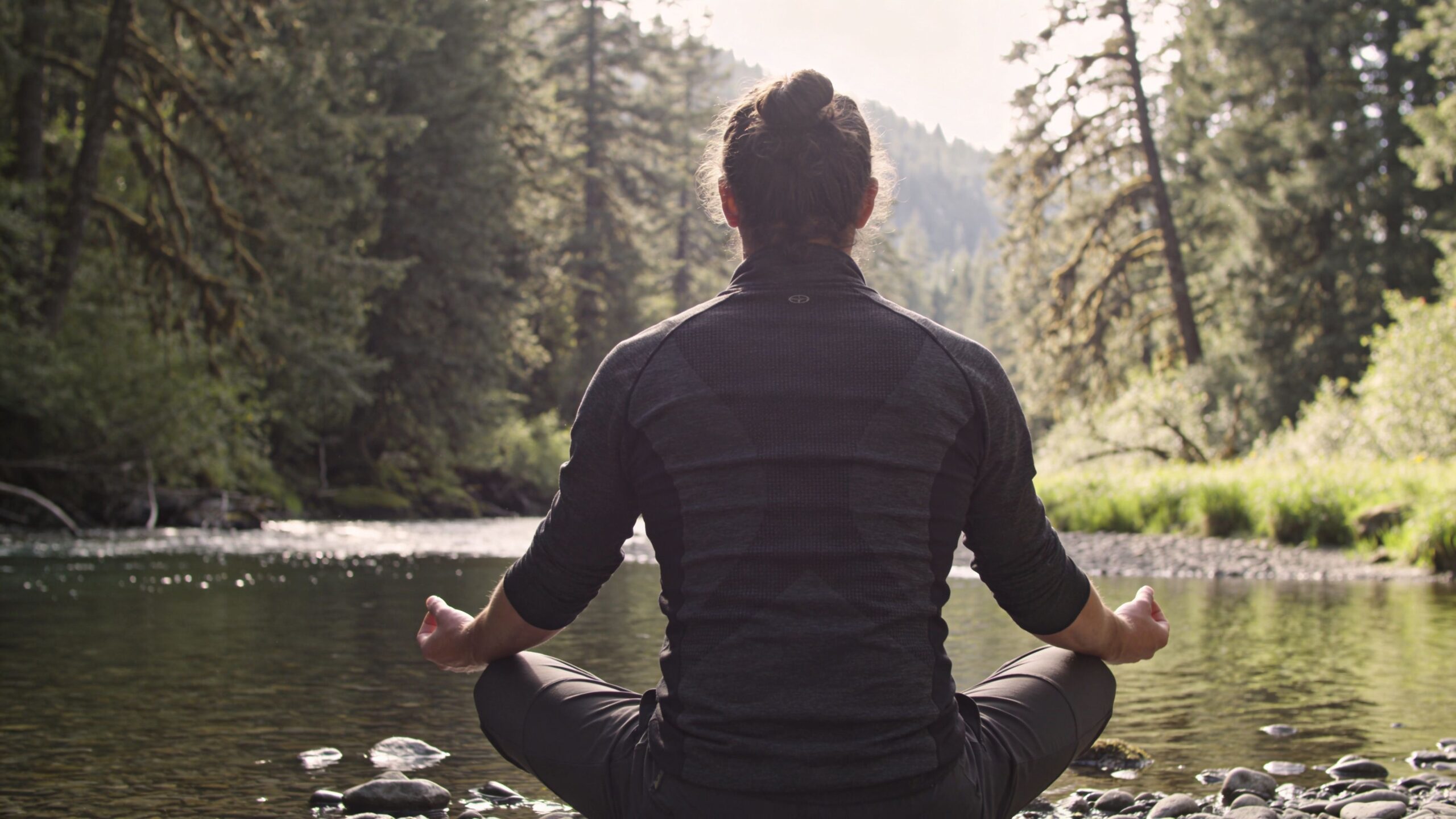 A man with a top knot meditating in a cross-legged position by a river in an Oregon forest.