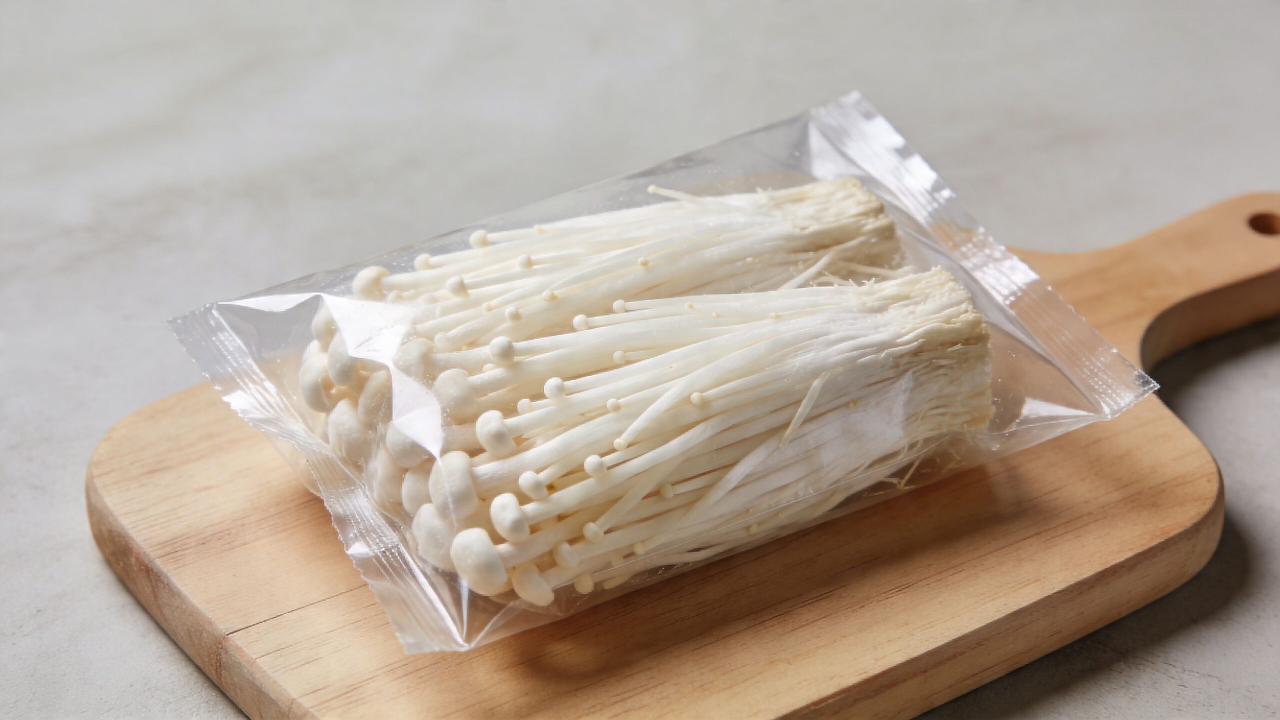 A pack of fresh white enoki mushrooms sitting on a wooden cutting board on a gray background.
