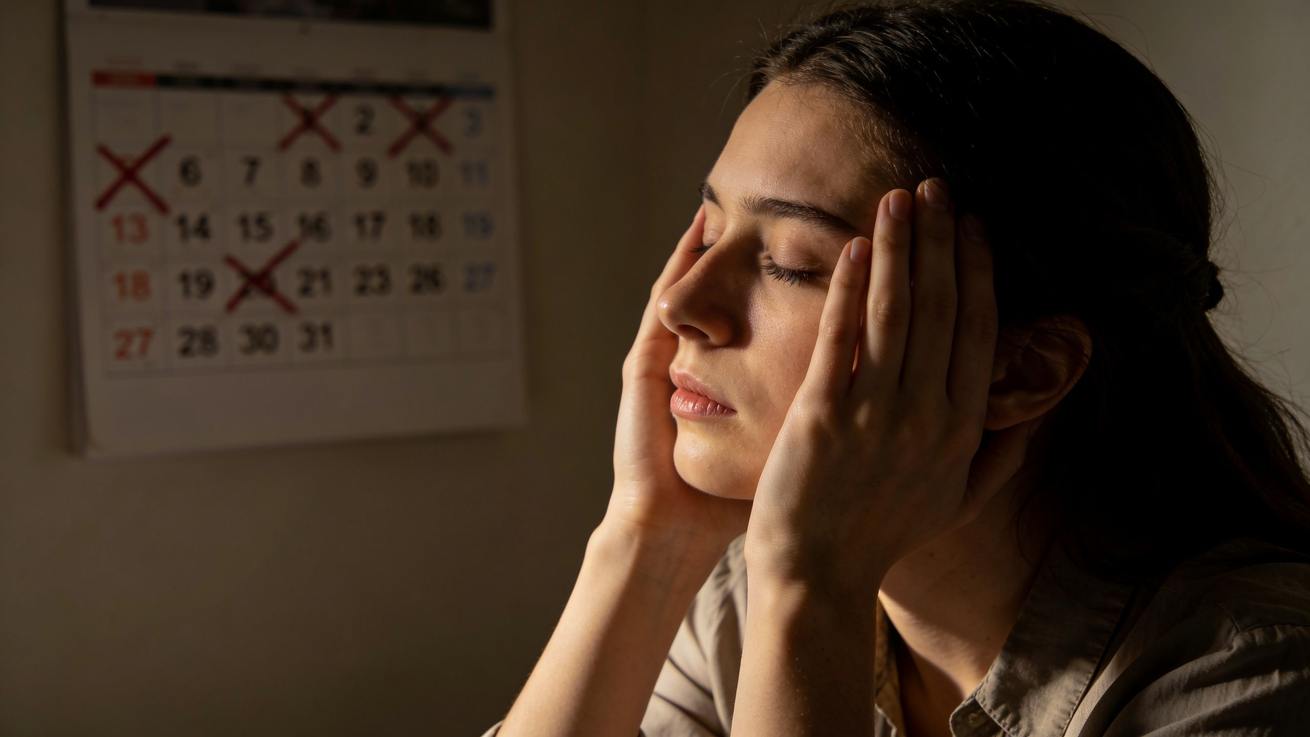 A stressed young woman holding her head while sitting in front of a marked calendar on the wall.